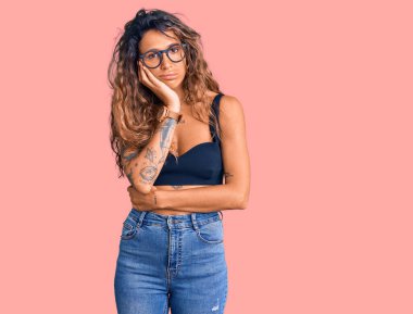 Günlük giysiler ve gözlük takan Latin kökenli genç bir kadın kollarını kavuşturarak depresyondan sıkılmış ve yorgun göründüğünü düşünüyor.. 