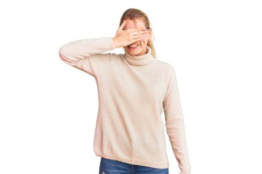 Balıkçı yaka kazak giyen, gözlüklü, gülümseyen ve gülümseyen genç, güzel sarışın kadın şaşırmak için gözlerini kapatıyor. kör kavram. 