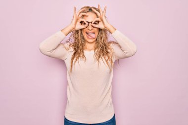 Genç, güzel, sarışın bir kadın pembe arka planda gözlük takıyor ve gözlük takıyor. Dürbün gibi jest yapıyor. Dürbün dilini çıkarıyor, gözler parmaklarından bakıyor. Çılgın ifade.