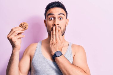 Sakallı, kolsuz tişörtlü, ağzı ağızla kaplı çikolatalı kurabiye yiyen, şok olmuş ve hata yapmaktan korkan genç yakışıklı bir adam. Şaşırtıcı ifade.