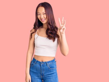 Genç, güzel Çinli kız, günlük kıyafetler giyip üç numaralı parmaklarıyla işaret ederken, kendinden emin ve mutlu bir şekilde gülümsüyor.. 