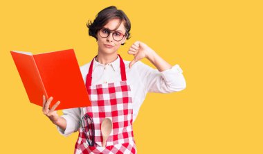 Kısa saçlı, profesyonel fırıncı önlüğü giyen, kızgın yüzlü yemek tarifi kitabı okuyan, olumsuz işaretli, baş parmakları aşağıda, reddedilme konsepti olan güzel bir genç kadın. 