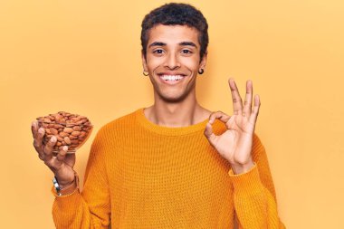 Genç Afrikalı Amerikalı adam elinde bademle kâse tutuyor tamam işareti yapıyor parmaklarıyla, gülümseyerek mükemmel bir sembol oluşturuyor 