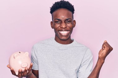 Genç Afrikalı Amerikalı adam domuz kumbarasını tutuyor gururla bağırıyor, zaferi kutluyor ve başarıyı kollarını kaldırarak kutluyor. 