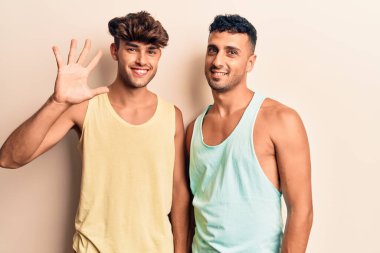 Genç eşcinsel çift gündelik kıyafetler giyer ve beş numarayı göstererek gülümserken kendinden emin ve mutlu. 