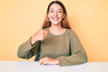 Genç esmer kadın, gündelik giysiler içinde masaya oturmuş mutlu ve pozitif gülümsüyor, başparmağını kaldırıyor, mükemmel ve onay işareti yapıyor. 