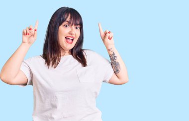 Genç ve cüsseli kadın gündelik kıyafetler giyiyor. Şaşırmış, şaşırmış, parmaklarıyla işaret ediyor ve kollarını kaldırıyordu.. 