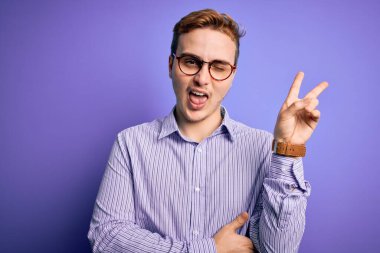Genç, kızıl saçlı, günlük gömlek ve gözlük takan, mor arka planda gülümseyen ve kameraya gülümseyen bir adam zafer işareti yapıyor. İki numara..