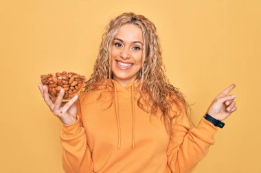 Sarı arka planda sağlıklı bademlerle kaseyi tutan genç, güzel sarışın kadın gülümsüyor.