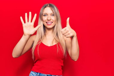 Genç sarışın kadın gündelik giysiler giyer ve altı numaralı parmaklarıyla işaret ederken kendinden emin ve mutlu bir şekilde gülümser.. 