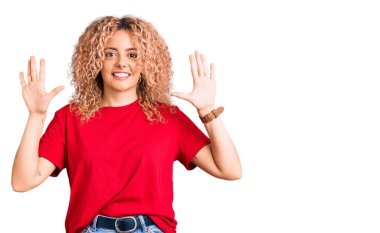 Kıvırcık saçlı, sıradan kırmızı tişörtlü, on numaralı parmaklarıyla onu gösteren, kendinden emin ve mutlu gülümseyen genç sarışın kadın.. 