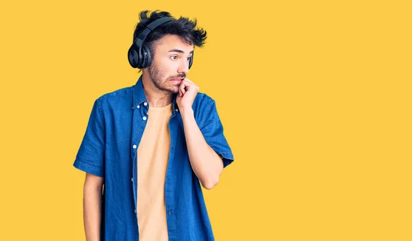 Young hispanic man listening to music using headphones looking stressed and nervous with hands on mouth biting nails. anxiety problem.