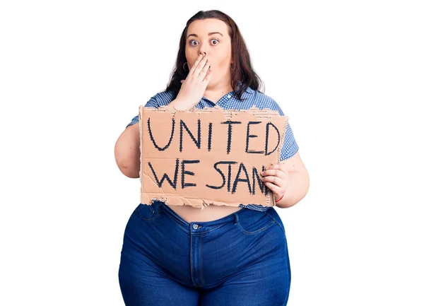 Young plus size woman holding united we stand banner covering mouth with hand, shocked and afraid for mistake. surprised expression