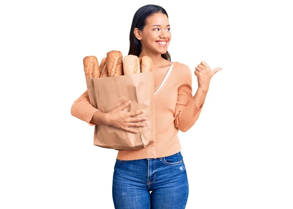 年轻美丽的拉丁女孩拿着纸袋 手拿着面包 大拇指朝外 笑容满面 张开嘴 — 图库照片