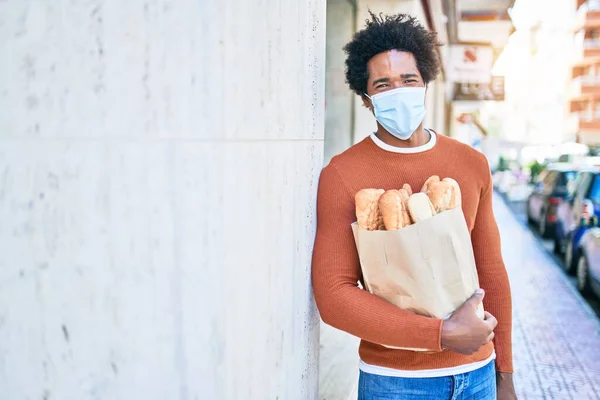 若いハンサムなアフリカ系アメリカ人の男性は医療マスクを着ている 街の通りでパンと紙の袋を保持壁にもたれる — ストック写真