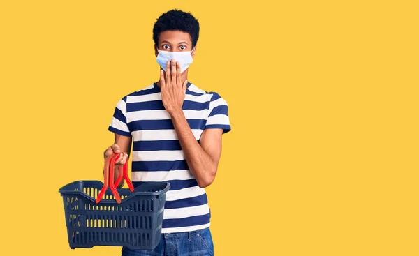 若いアフリカ系アメリカ人の男性がショッピングバスケットや医療用マスクを手で口をカバーし ショックを受け ミスを恐れている 驚きの表情 — ストック写真
