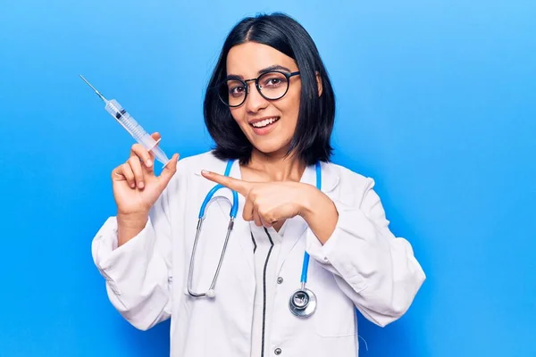 Young beautiful latin woman wearing doctor stethoscope holding syringe smiling happy pointing with hand and finger