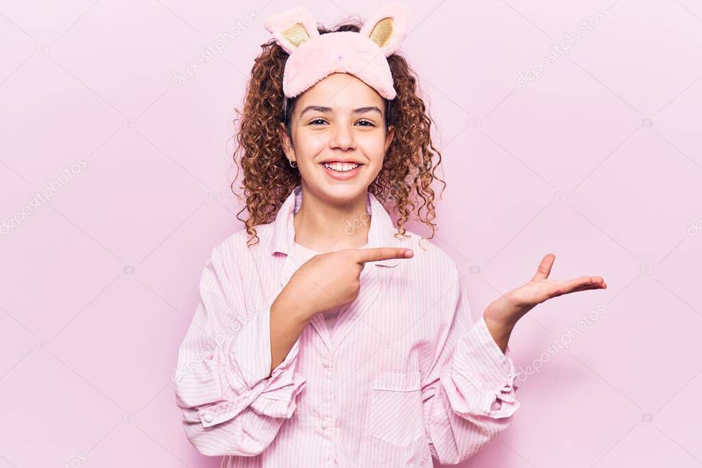 Beautiful kid girl with curly hair wearing sleep mask and pajamas amazed and smiling to the camera while presenting with hand and pointing with finger. 