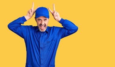 Orta yaşlı, tamirci üniforması giyen, komik ve deli pozu veren, parmakları tavşan kulaklı, neşeli gülümseyen bir adam. 