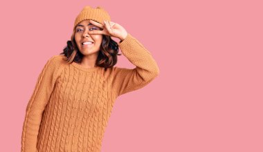 Genç, güzel, melez bir kadın yün kazak ve kış şapkası giyiyor. Barış sembolü yapıyor. Yüzünde parmakları var. 