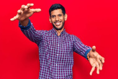 Gündelik giysiler giyen genç Latin adam kameraya bakıp sarılmak için kollarını açmış gülümsüyor. Mutluluğu kucaklayan neşeli bir ifade.. 