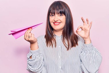 Elinde kağıt uçak tutan genç ve cüsseli bir kadın parmaklarıyla imza atıyor, gülümseyerek mükemmel bir işaret veriyor. 