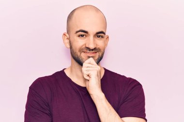 Genç, kel, sıradan bir tişört giyen, kolları çapraz ve çenesi açık kameraya gülümseyen yakışıklı bir adam. Olumlu düşünmek. 