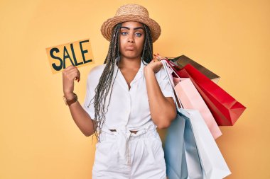 Saç örgülü, alışveriş torbası ve satış afişi taşıyan genç Afro-Amerikalı kadın depresif ve endişeli, sinirli ve korkmuş. hüzünlü ifade. 