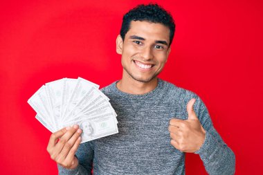 Genç, yakışıklı, elinde dolar sembolü olan para çantası tutan mutlu ve pozitif gülümseyen, başparmağıyla mükemmel ve onay işareti yapan bir İspanyol. 
