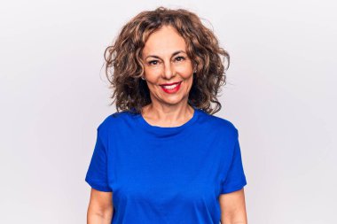 Orta yaşlı, esmer, mavi tişörtlü güzel bir kadın beyaz arka planda mutlu ve havalı bir gülümsemeyle dikiliyor. Şanslı kişi..