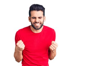 Sakallı, sıradan tişörtler giyen, çok mutlu ve heyecanlı genç bir adam kolları havada, gülümseyen ve başarı için çığlık atan, kazanan bir jest yapıyor. kutlama konsepti. 