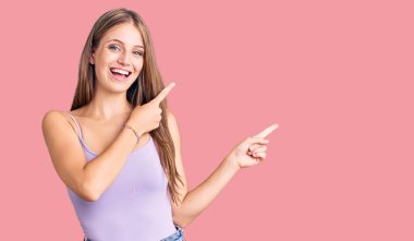 Kolsuz tişörtlü, sıradan giyinen genç, güzel sarışın kadın gülümseyip kameraya bakıyor ve iki eliyle ve parmaklarıyla yan tarafı işaret ediyor.. 