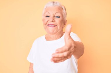 Son sınıf güzeli, mavi gözlü, gri saçlı, sarı arka planda klasik beyaz tişört giyen, dostça bir selamlama ve hoş geldin hediyesi olarak elini sıkan bir kadın. başarılı iş. 