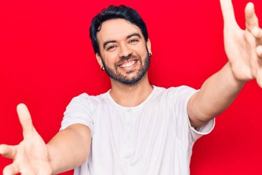 Gündelik giysiler giymiş genç İspanyol adam kameraya bakıp sarılmak için kollarını açmış gülümsüyor. Mutluluğu kucaklayan neşeli bir ifade.. 