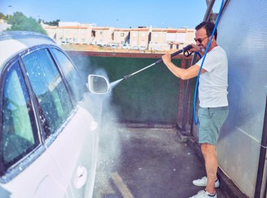 Orta yaşlı yakışıklı, günlük kıyafetler giyen ve mutlu gülümseyen güneş gözlüklü bir adam. Yüzünde gülümsemeyle ayakta durup su tabancası kullanarak araba yıkamak..