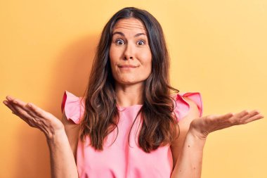 Genç, güzel esmer, pembe günlük tişört giyen, sarı arka planda duran, kolları açık, hiçbir fikri olmayan, şüpheli yüzlü bir kadın..
