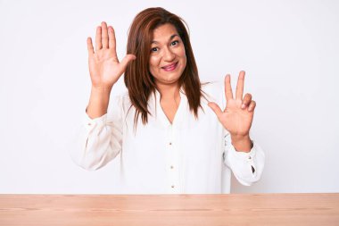Orta yaşlı esmer, İspanyol iş kadını. Masada oturan ve 8 numaralı parmaklarıyla işaret eden, kendinden emin ve mutlu bir şekilde gülümseyen gündelik beyaz gömlekli kadın.. 
