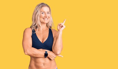 Orta yaşlı, sarışın, bikinili, yüzünde kocaman bir gülümseme olan, elleri ve parmaklarıyla kameraya bakan bir kadın.. 