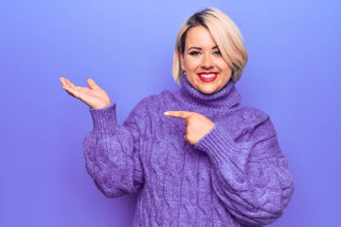 Güzel sarışın ve boyunda bir kadın mor arka planda balıkçı yaka kazak giyiyor. Kameraya hayret ediyor ve gülümsüyor. Elle gösterip parmağıyla işaret ediyor..