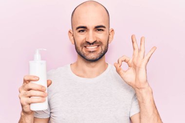Genç yakışıklı kel adam elinde krem losyon şişesi tutuyor. Parmaklarıyla imza atıyor, gülümseyerek mükemmel bir sembol oluşturuyor. 