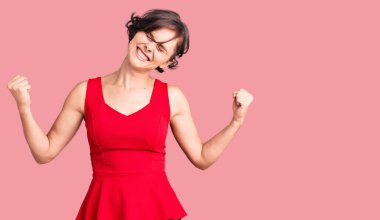 Kısa saçlı, kolsuz tişörtlü, rahat giyinen çok mutlu ve heyecanlı genç bir kadın kolları havada, gülümseyerek ve başarı için çığlık atarak zafer hareketi yapıyor. kutlama konsepti. 