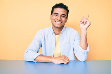 Genç, yakışıklı, günlük kıyafetler giyen, gülümseyen, elleri ve parmakları yanda mutlu mesut oturan İspanyol bir adam. 