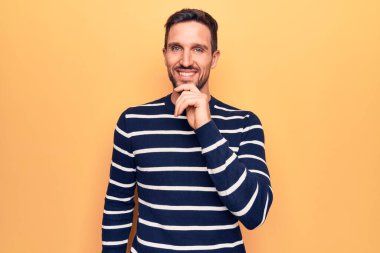 İzole edilmiş sarı arka plan üzerinde duran sıradan çizgili süveter giyen genç ve yakışıklı adam kameraya kollarını kavuşturup çenesine dokunarak gülümsüyor. Olumlu düşünüyorum..