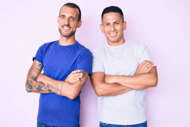 Sıradan kıyafetler giyen, mutlu yüzlü, kollarını kavuşturup kameraya bakan iki genç gay. pozitif kişi. 