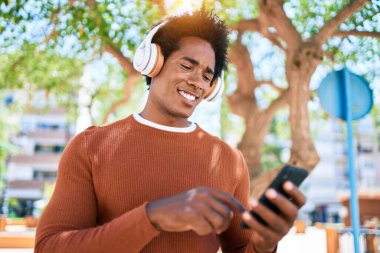 Genç, yakışıklı, kulaklık takan Afrikalı Amerikalı bir adam. Yüzünde gülücükle, akıllı telefon kullanarak şehrin caddesinde yürüyorsun..