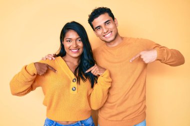 Güzel, Latin, genç bir çift, günlük giysiler giyip, kendilerine güveniyorlar, gülümsüyorlar, parmaklarını gururla ve mutlu bir şekilde işaret ediyorlar.. 
