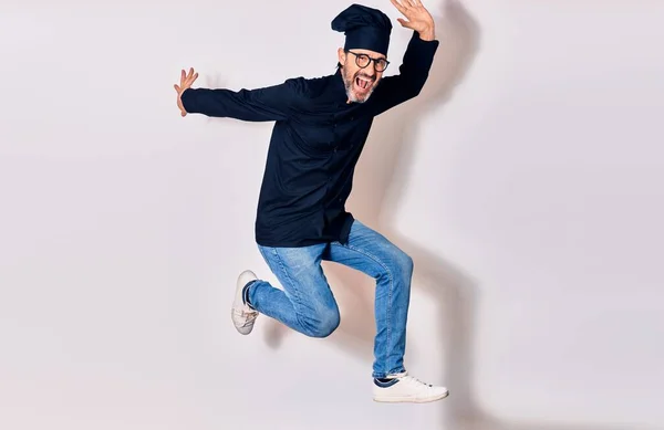 Middle age handsome hispanic man wearing glasses and cook uniform smiling happy. Jumping with smile on face over isolated white background