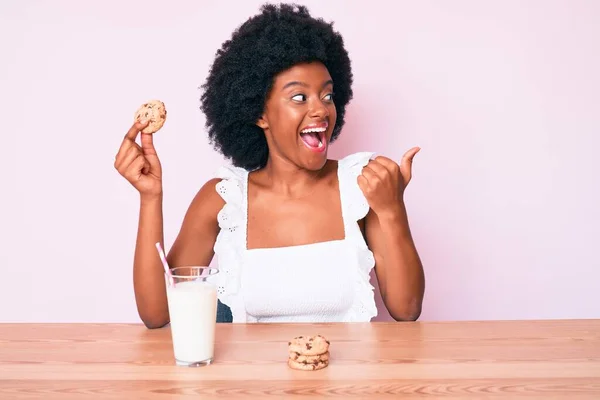 Young african american woman drinking glass of milk holding cookie pointing thumb up to the side smiling happy with open mouth