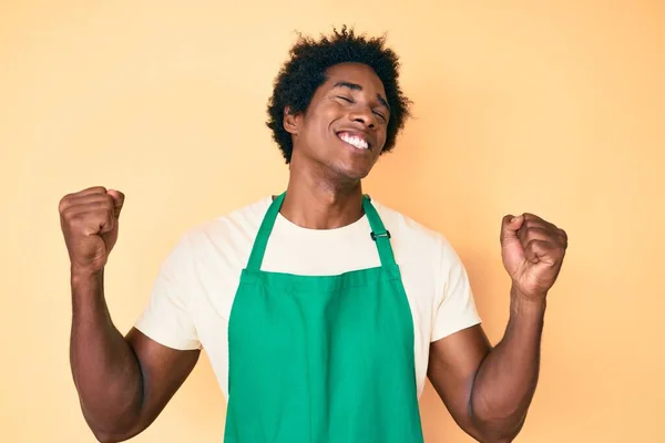 Handsome african american man with afro hair wearing waiter apron very happy and excited doing winner gesture with arms raised, smiling and screaming for success. celebration concept.