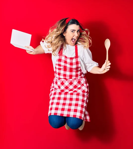 Young beautiful blonde cook woman wearing apron smiling happy. Jumping with smile on face holding recipe book and spoon over isolated red background